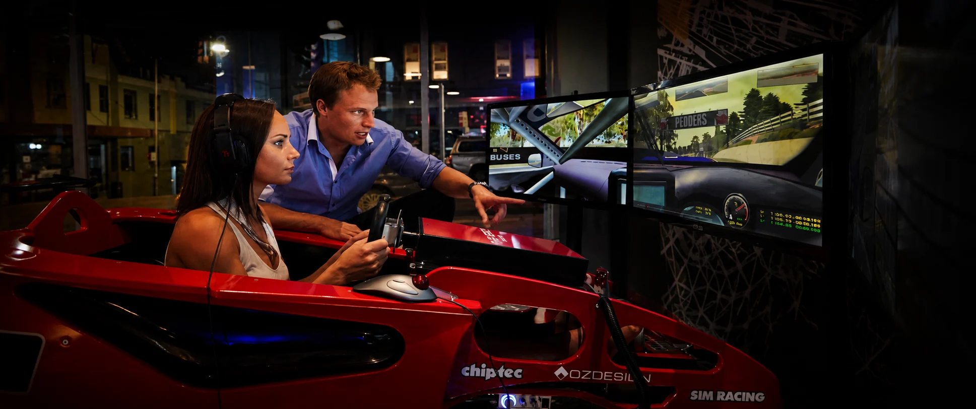 An Excape coach guides a guest at the wheel of a red simulator in a street-front sim racing space